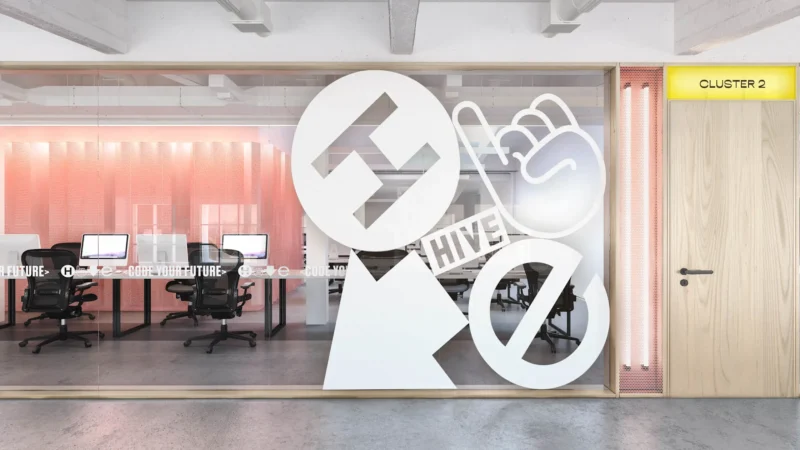 A modern office interior at Hive Helsinki, seen through a glass partition with prominent white vinyl graphics featuring the Hive logo, a pointing finger, and other abstract shapes. Inside, there are desks with computers and office chairs, and the wall behind them is a translucent, textured screen in a pinkish hue. A wooden door to the right of the glass reads "CLUSTER 2" on a yellow sign above it.