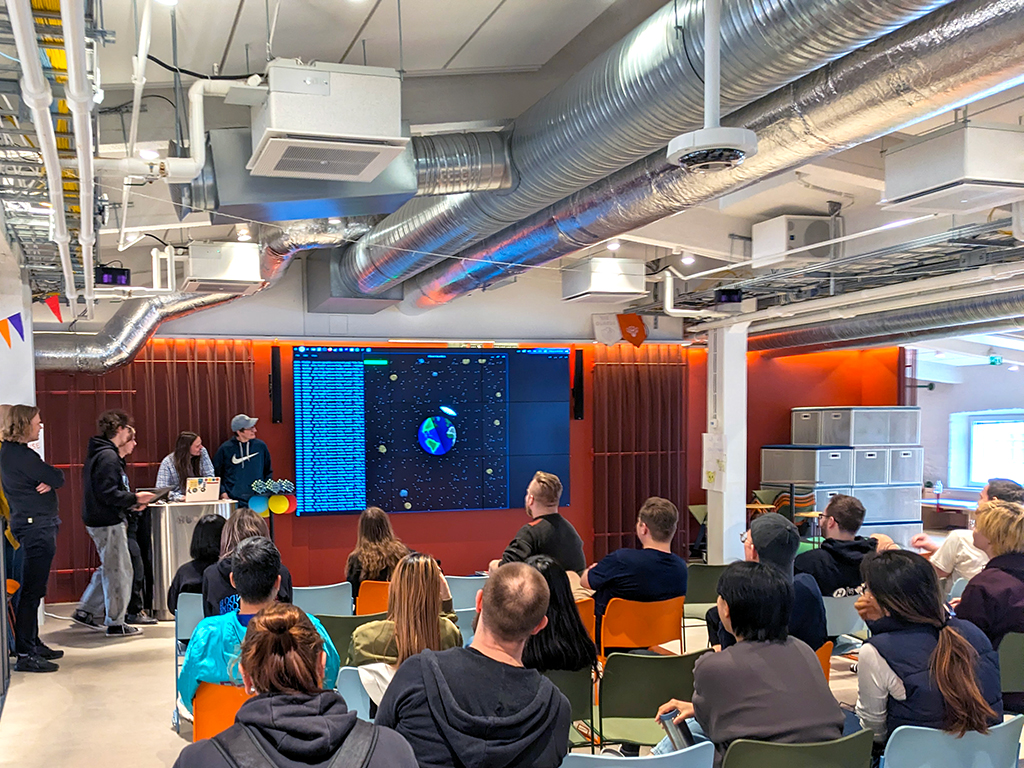 An engaging presentation at Hive Helsinki, during the Game Jam 2025 event. A large screen displays what appears to be game code and a 3D graphic of planets in space. Several people are gathered near the screen, possibly presenters or participants, while a seated audience watches from colorful chairs. The room has an industrial aesthetic with exposed ceiling ducts.
