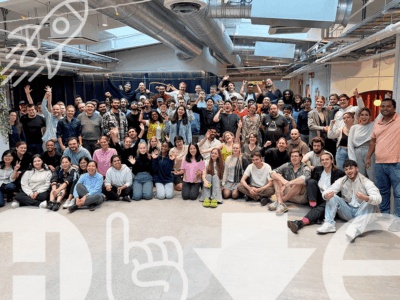 A large and diverse group of smiling individuals, appearing to be the vibrant community of Hive Helsinki, posing for a group photo in their modern, open-plan learning space. Many are waving enthusiastically, showcasing the energy and collaborative spirit of the "Road to Futureproofing Finland" initiative. Overlaid transparent graphics include a rocket ship, the Hive logo, and an arrow, symbolizing progress and innovation.