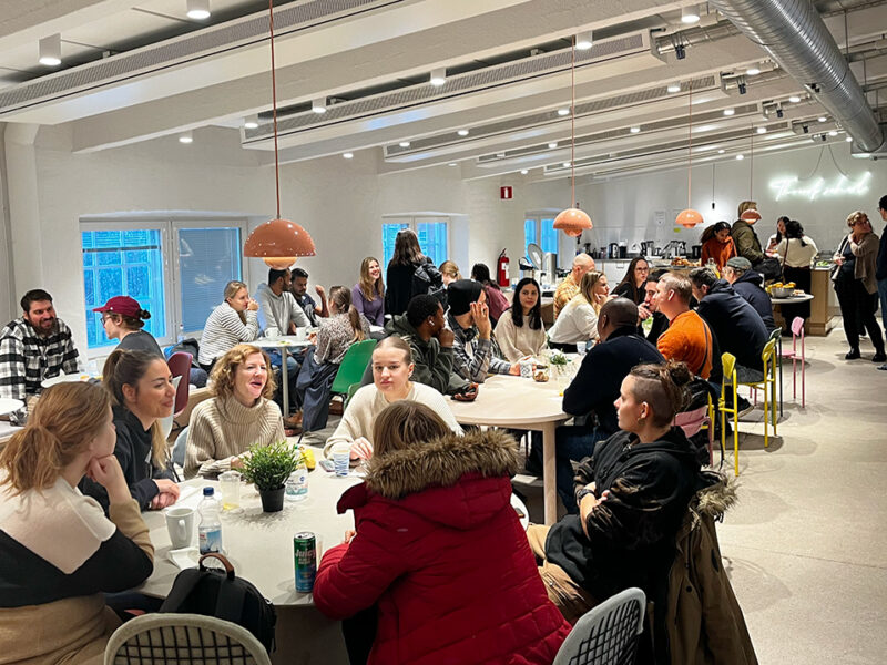 A lively and engaging gathering of a diverse group of Hive Helsinki students and community members in a spacious common area or cafeteria. People are seated at various tables, conversing and enjoying refreshments, highlighting the vibrant campus life and opportunities for networking and collaboration at Hive Helsinki. The bright and modern interior, located in Helsinki, Finland, fosters a supportive environment for tech education and futureproofing skills.