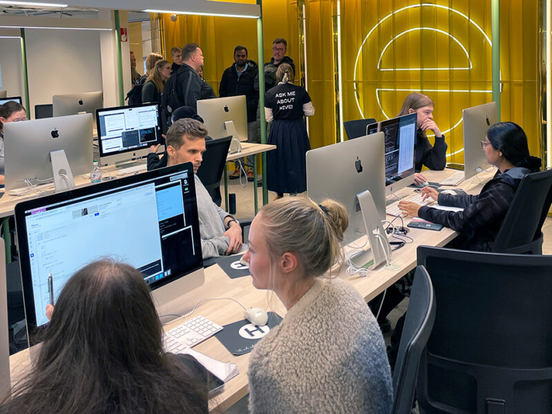 Students and visitors actively engaged in a coding or learning session within the bright, modern computer lab at Hive Helsinki. Individuals are seated at desks, focused on their Apple iMac computers, while others stand and observe, demonstrating a collaborative learning environment. A person in the background wears a shirt that says "ASK ME ABOUT HIVE!", highlighting the open and informative atmosphere. The space features striking yellow wall panels and a large illuminated 'E' graphic.