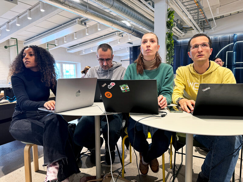 Four adult students or participants intently focused on their laptops at a collaborative workspace within Hive Helsinki. They are engaged in deep work or a coding challenge, highlighting the immersive and practical learning environment for futureproofing careers in Finland. The modern campus features industrial-style ceilings and bright lighting, conducive to peer-to-peer learning.