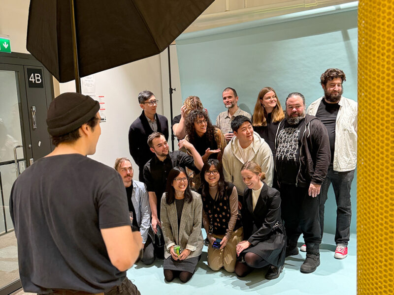A group of diverse individuals, likely Hive Helsinki students or community members, posing for a professional photo shoot with a light blue backdrop. A photographer is visible in the foreground, capturing the moment. The group includes people standing and kneeling, showcasing a friendly and collaborative tech education environment focused on futureproofing skills in Finland.