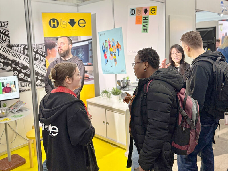 A vibrant Hive Helsinki booth or stand at an event, where staff members are engaging with potential students or visitors. In the foreground, a person in a "Hive" branded hoodie is speaking with another individual, while in the background, a person with a beard stands near "Hive" branded banners and a screen showing content. This scene showcases Hive Helsinki's active presence in promoting coding education and futureproofing skills in Finland.