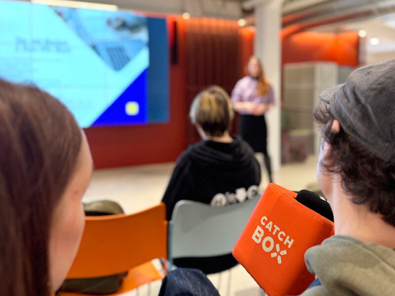 An interactive presentation in progress at Hive Helsinki, where an audience member in the foreground holds an orange 'CATCH BOX' microphone, indicating a Q&A session. A speaker is visible at the front, presenting on a large screen. This captures the engaging and collaborative atmosphere of coding workshops and tech education events at the Hive Helsinki campus in Finland, fostering futureproofing skills.