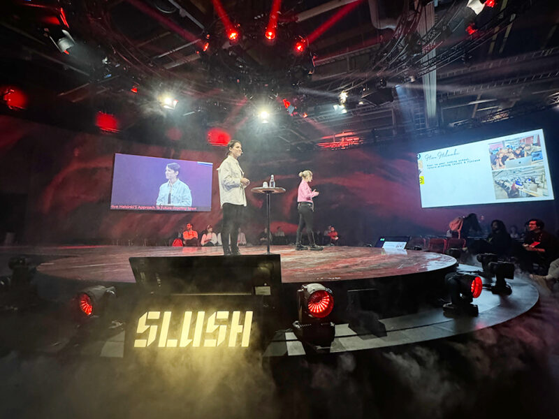 A dynamic presentation stage at Slush, a major startup and tech event in Helsinki, featuring two speakers presenting with large screens displaying content. The stage is enveloped in fog with illuminated "SLUSH" lettering. This image captures Hive Helsinki's presence at prominent Finnish tech events, showcasing their role in futureproofing talent and coding education in the startup ecosystem.