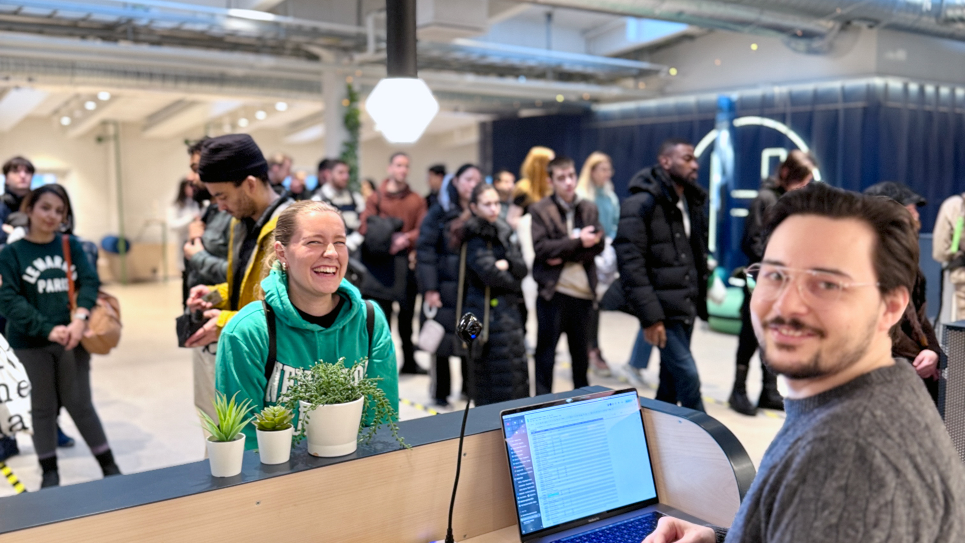 A lively scene at the registration desk for a "Piscine" event at Hive Helsinki. A person in a green hoodie is laughing happily at the counter, where small potted plants are also visible. In the foreground, a person with glasses smiles at the camera from behind a laptop. A queue of diverse individuals is seen in the background, waiting for entry.