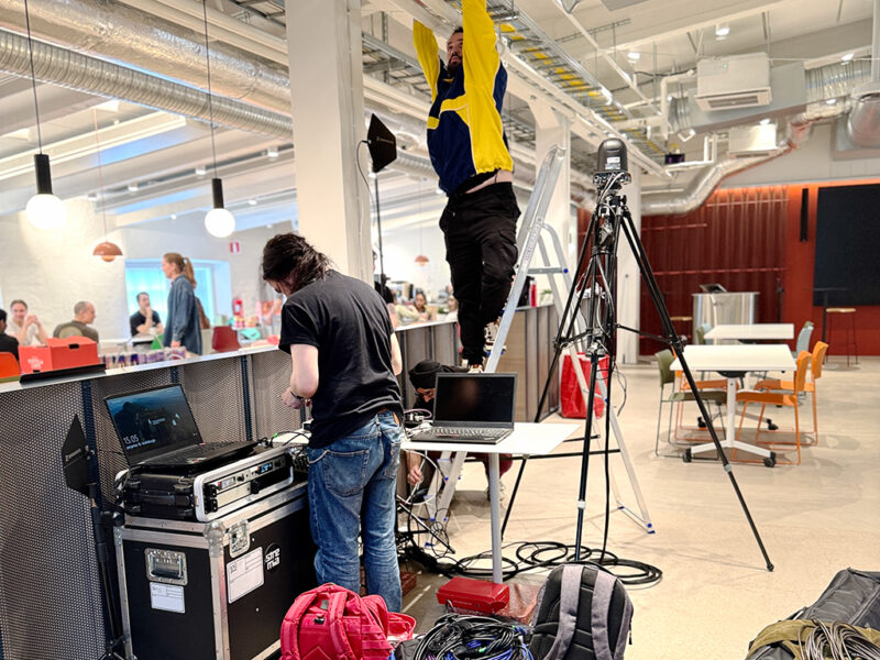 Two individuals setting up technical equipment in a spacious indoor venue, likely for an event. One person is on a ladder adjusting overhead cables, while another is at a table with laptops and audio-visual gear in a rack case, connecting cables. The background shows a modern space with tables and chairs, and other people in the distance.