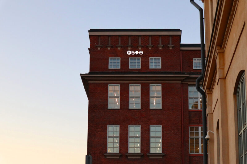 Exterior view of a red brick building, possibly Hive Helsinki at Haapaniemenkatu 5, with white-framed windows. The building features modern white signage with abstract symbols visible on the upper facade against a clear sky.