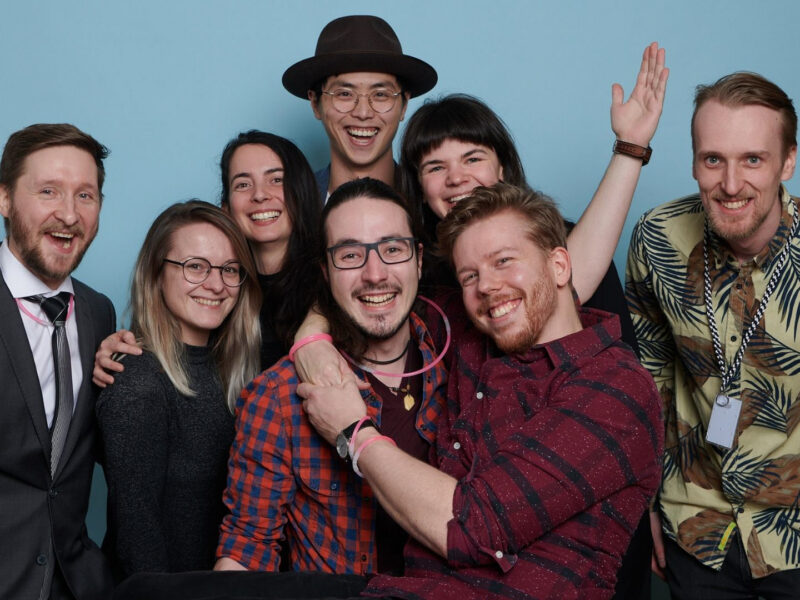 A group of eight diverse young adults smiling and posing closely together in front of a blue background, showing excitement and camaraderie.