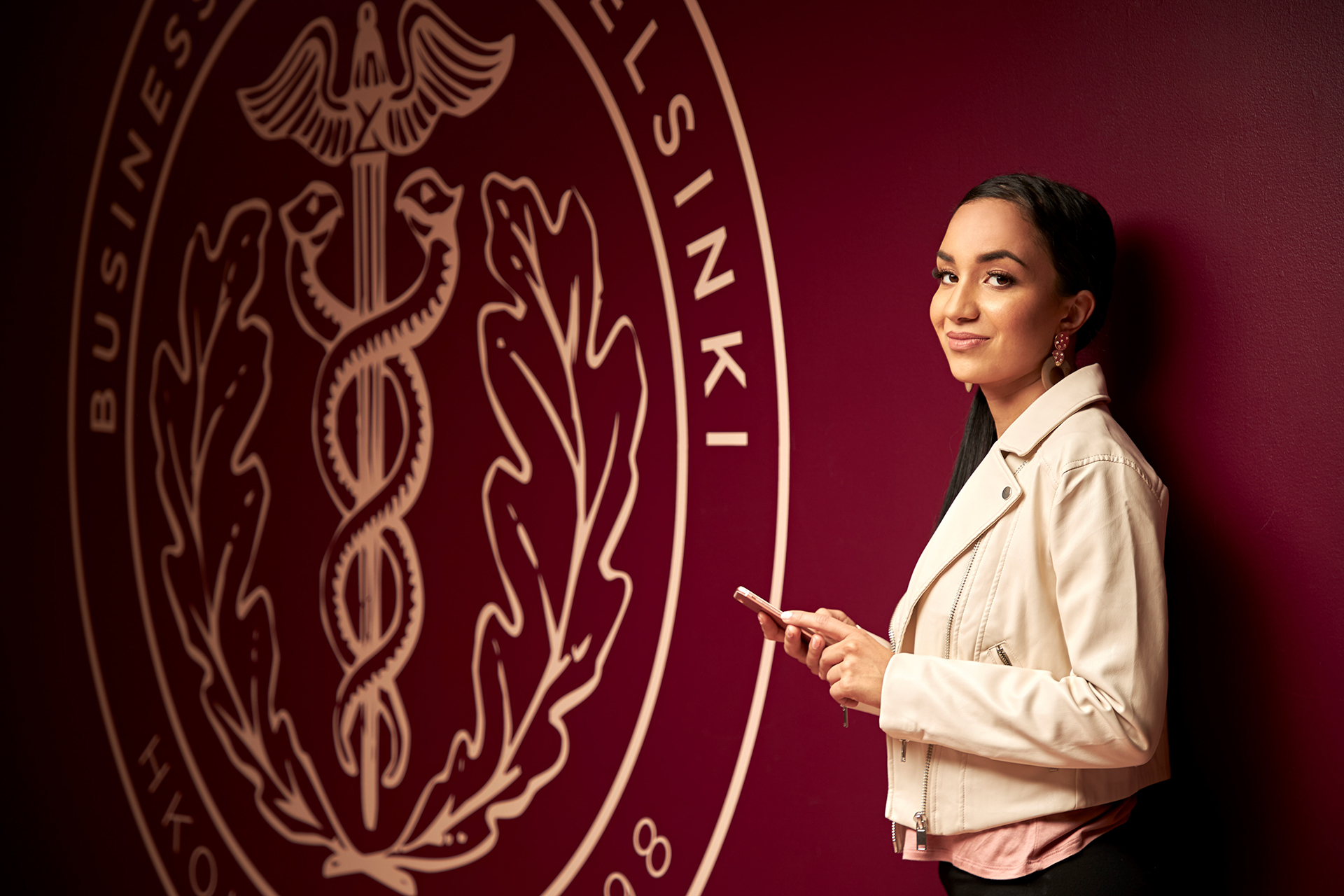 A smiling young woman, standing next to a large, prominent logo of Business College Helsinki on a dark red wall. She is wearing a light-colored jacket over a dark top and holding a smartphone, looking towards the viewer.