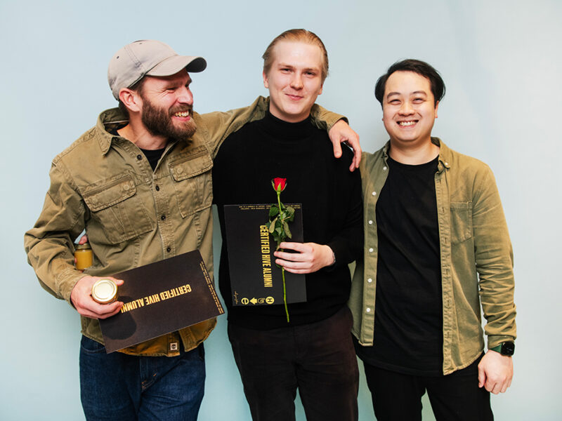 Three smiling male alumni of Hive Helsinki posing together, with one holding a "CERTIFIED HIVE ALUMNI" certificate and a red rose. They appear to be celebrating their achievements, symbolizing the strong community and successful outcomes of the tech education program in Helsinki.