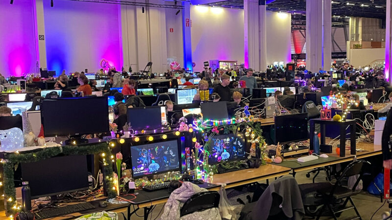 A large hall at the Assembly LAN party event in Helsinki, filled with rows of computer stations where individuals are intensely gaming. Many setups are adorned with colorful lights and decorations, showcasing the personal flair and dedication of the gaming community, often including Hive Helsinki students.