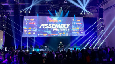 A large stage at the Assembly Winter 2024 event in Helsinki, with a massive screen displaying "Welcome to ASSEMBLY WINTER '24" and various sponsor logos. Bright stage lights illuminate the area, where a speaker addresses a large audience, highlighting Hive Helsinki's connection to Finland's vibrant tech and gaming community.