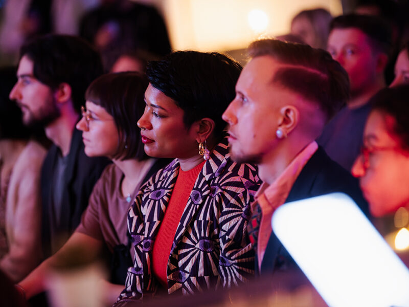 A diverse group of attendees, primarily young adults, seated and attentively watching a presentation or event at Hive Helsinki. The focus is on a woman in the foreground with dark hair and a patterned jacket, showcasing the inclusive and engaging atmosphere of a tech education community in Helsinki.