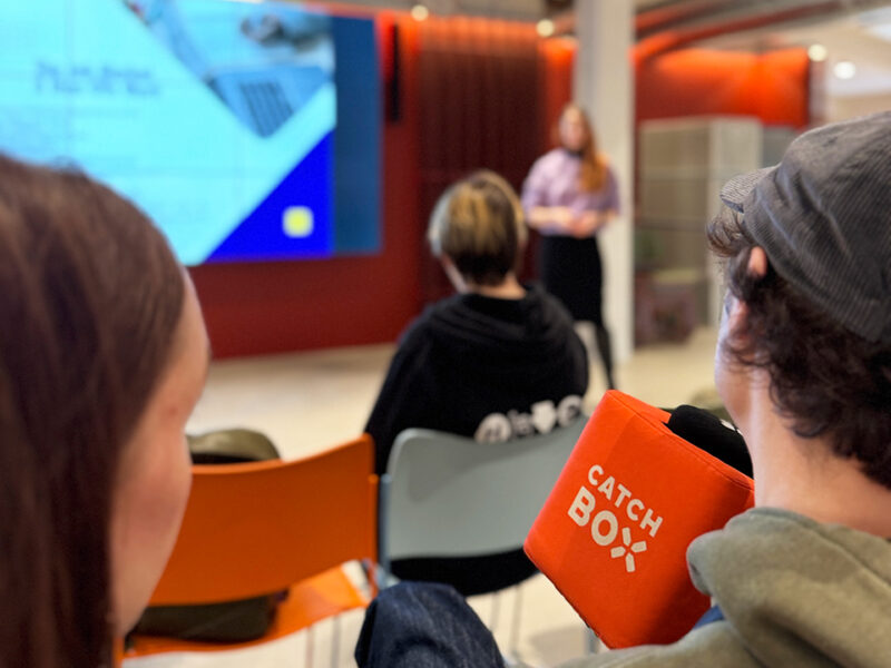 An audience member, seen from behind, holding an orange "CATCH BOX" microphone cube, indicating an interactive Q&A session. A presenter is visible on stage in the background, speaking in front of a large screen. Other audience members are also visible, engaged in the presentation in a modern event space at Hive Helsinki.