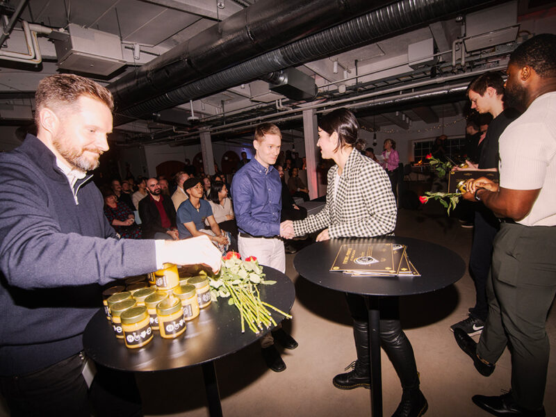 An award ceremony or graduation event at Hive Helsinki, where a man and a woman are shaking hands across a table with award certificates and red roses. Jars of honey or a similar gift are also visible on the table. In the background, an audience is seated, indicating a celebratory moment for Hive Helsinki graduates or partners.