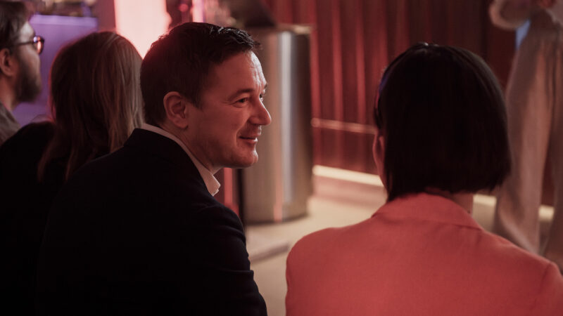 A close-up of a man smiling and looking at another attendee during a Hive Helsinki event. The soft, red ambient lighting creates a warm and intimate setting, showcasing the friendly interactions and community building that takes place at our tech education gatherings in Helsinki, Finland.