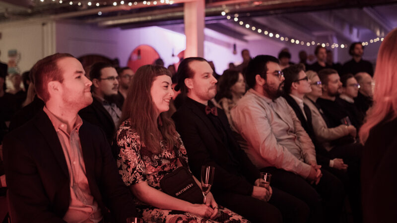 A row of diverse attendees at a Hive Helsinki event, seated and attentively watching a presentation. Many are smiling and engaged, with string lights visible in the background, highlighting the interactive and inspiring environment for learning and networking within the Helsinki tech community.