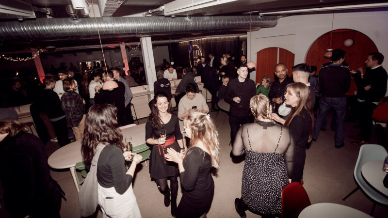 A bustling scene at a Hive Helsinki event, with numerous guests mingling, chatting, and holding drinks. The modern venue features exposed industrial elements and warm lighting, emphasizing the vibrant social opportunities for students and professionals in the Helsinki tech ecosystem.
