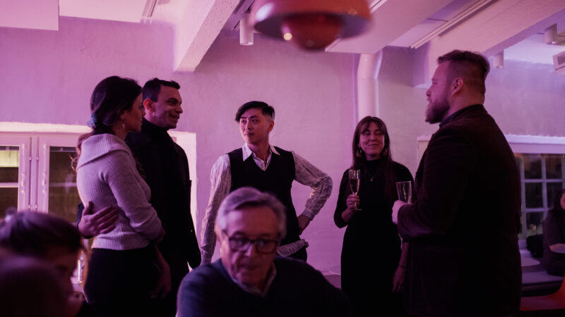 A diverse group of people socializing and conversing at a Hive Helsinki event, illuminated by purple ambient lighting. Individuals are holding drinks and engaging in lively discussions, showcasing the welcoming and dynamic atmosphere for networking within the tech community in Helsinki, Finland.
