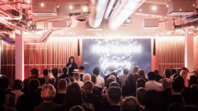 A speaker on stage at Hive Helsinki, addressing a large audience in a spacious event hall. The prominent screen behind the speaker displays "Hive Helsinki" with an abstract pattern. The room is dramatically lit with red tones and features exposed industrial ductwork, setting the scene for an engaging tech education presentation in Helsinki, Finland.