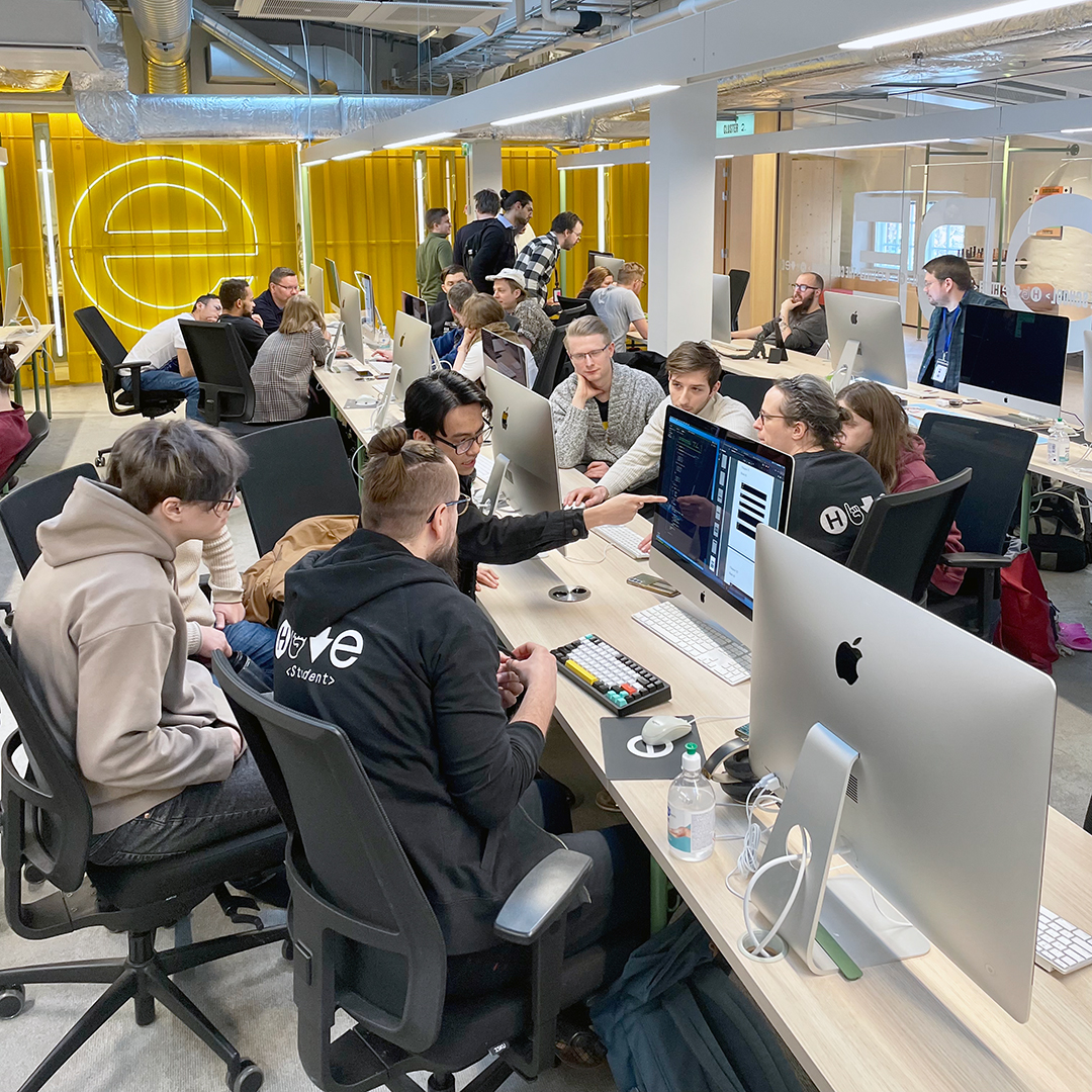 An active and collaborative open-plan workspace at Hive Helsinki, filled with people working on Apple iMac computers. In the foreground, a group is gathered around a desk, with one person pointing at a screen that displays code or a programming interface, indicating teamwork and problem-solving. The background features bright yellow panels and a neon sign.