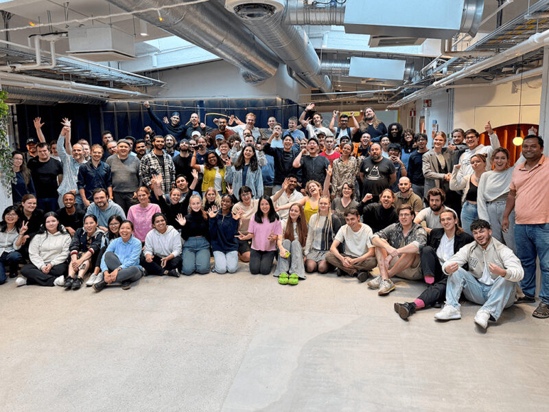 A large, diverse group of people, appearing to be students and staff of Hive Helsinki, gathered for a group photo in a bright, open-plan space. Many are smiling and waving, creating a cheerful and energetic atmosphere. Some are standing, while others are seated or kneeling in the front.