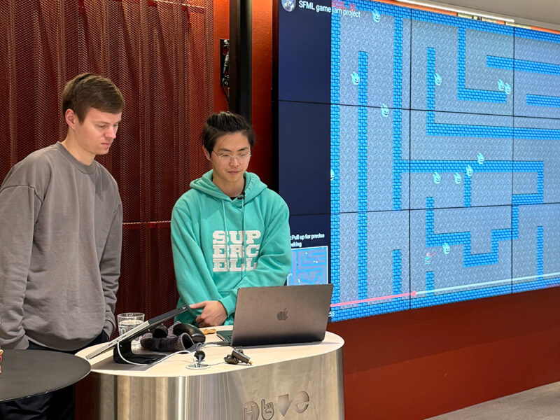 Two Hive Helsinki students presenting a game project at Demoday. One student wears a Supercell hoodie, and they stand in front of a large screen displaying a retro-style game map and code. This showcases practical coding skills and project-based learning at the Hive Helsinki campus in Finland.