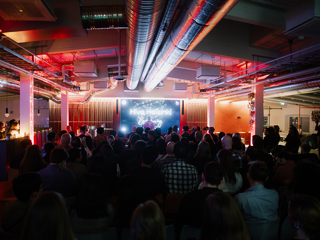 A wide shot of a dark, packed auditorium at Hive Helsinki during an event, with an audience facing a large screen displaying "Hive Helsinki" and content. Red ambient lighting illuminates the stage and industrial ceiling pipes, highlighting an engaging atmosphere for a tech or coding presentation in Helsinki.