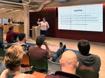 A speaker presents "Job hunting for realists" to an audience in a modern event space in Hive Helsinki. The audience is seated, attentively listening, with some visible from behind.