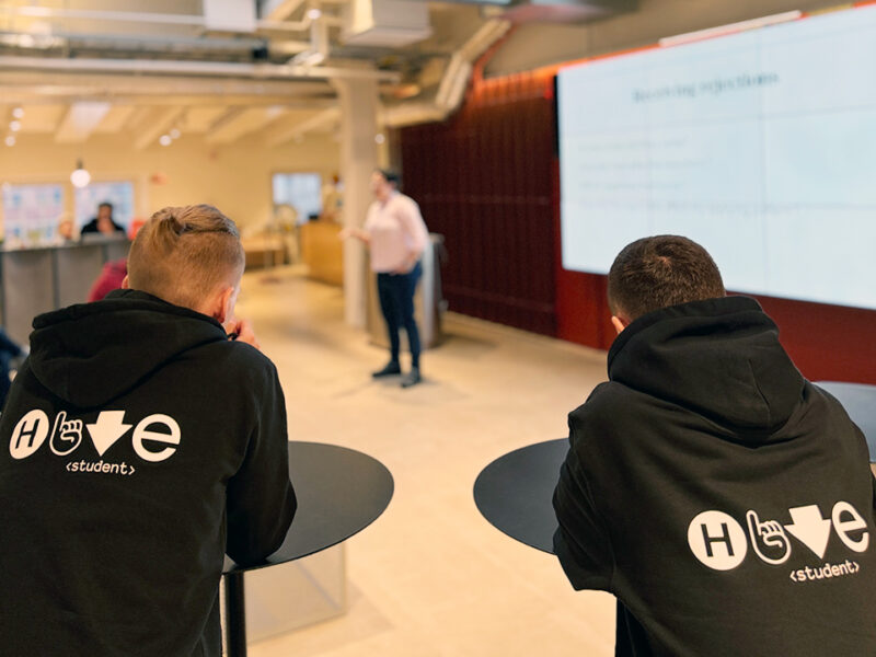 A speaker presents "Job hunting for realists" to an audience in a modern event space in Hive Helsinki. The audience is seated, attentively listening, with some visible from behind.