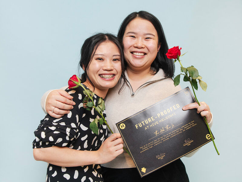 Two smiling female graduates from Hive Helsinki embracing and proudly holding their "FUTURE-PROOFED AT HIVE HELSINKI" certificates and red roses. They stand against a light blue background, symbolizing the success and job readiness achieved through their **coding education** in **Helsinki**, preparing them for **futureproofing careers**.