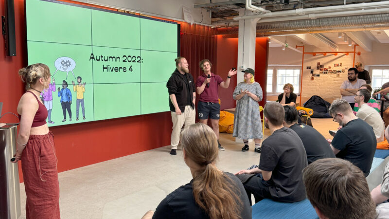 A group of Hive Helsinki students presenting "Autumn 2022: Hivers 4" to an audience in a vibrant event space. The screen displays an illustration of people with thought bubbles containing gears, symbolizing collaborative problem-solving and project-based learning at the coding school in Helsinki, Finland.
