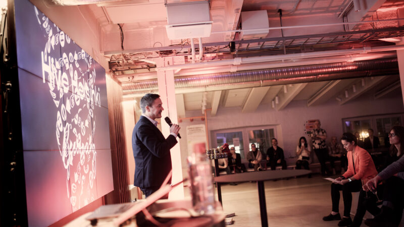 Ilkka Paananen, a speaker at a Hive Helsinki event, stands on stage holding a microphone in front of a large screen displaying abstract white text. An audience is visible in the background, listening attentively, showcasing the engaging presentations and community events held at the tech education campus in Helsinki, Finland.