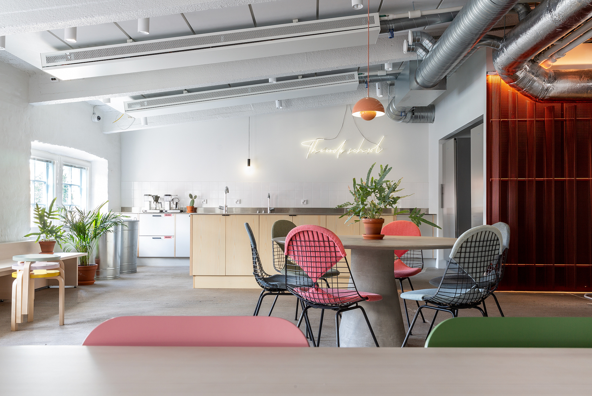 A bright and modern kitchen and lounge area at Hive Helsinki, featuring light wood cabinetry, a large island, and various plants. A round table with colorful chairs, including pink and grey, is in the foreground. In the background, there's a white tiled wall with a neon sign that says "The code school". Exposed ductwork is visible on the ceiling.
