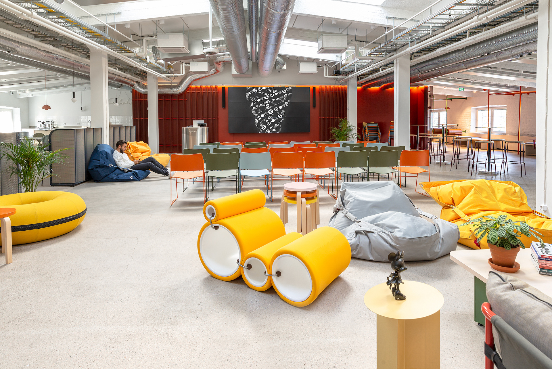 A spacious, modern lounge and auditorium area at Hive Helsinki, featuring a large screen at the back, rows of colorful chairs, and various casual seating options including bean bags and unique yellow tubular seating elements. Exposed ductwork and an industrial aesthetic are visible, with a person relaxing on a blue bean bag in the background.