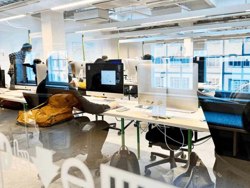 An interior view of a modern computer lab or open-plan workspace at Hive Helsinki, with rows of Apple iMac computers on light-colored desks. Several individuals are seated and working, reflected in a glass partition in the foreground. The room is brightly lit with an industrial-style ceiling and large windows in the background.