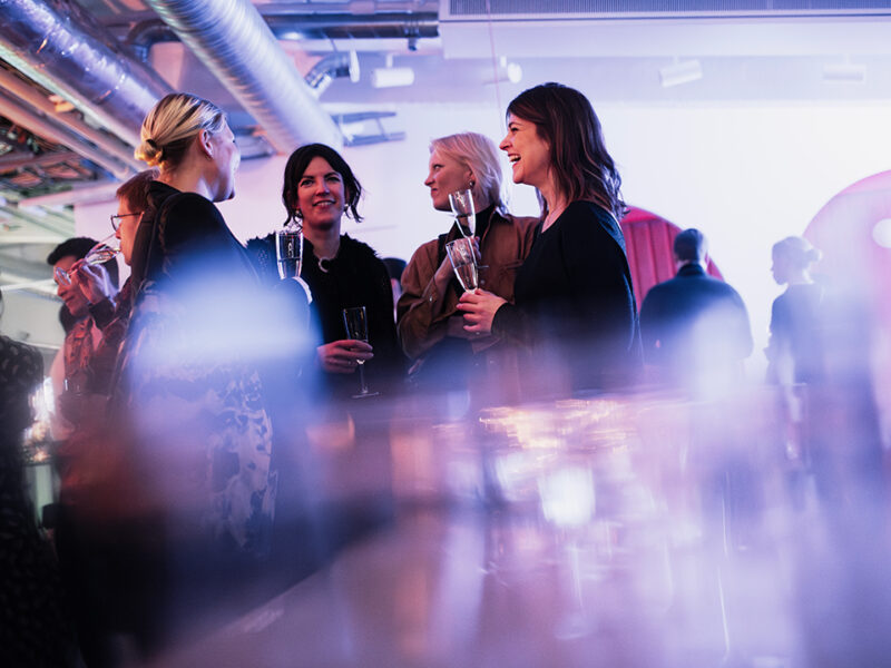 A group of smiling individuals, mostly women, chatting and holding champagne glasses at a lively networking event at Hive Helsinki. The foreground is blurred with lights, creating a festive atmosphere for guests at a launch or celebration in Helsinki.