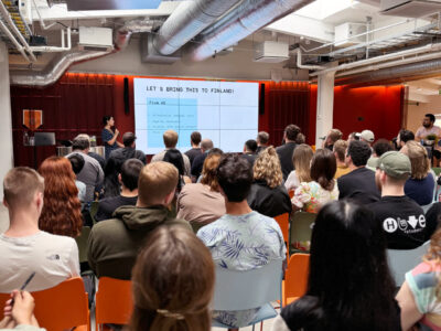 A speaker presenting to a large audience at Hive Helsinki, with a screen displaying "LET'S BRING THIS TO FINLAND!" and bullet points about attitude, work-life balance, and vocational concepts. This highlights Hive Helsinki's mission to foster tech talent and innovation in Finland through its unique coding education.