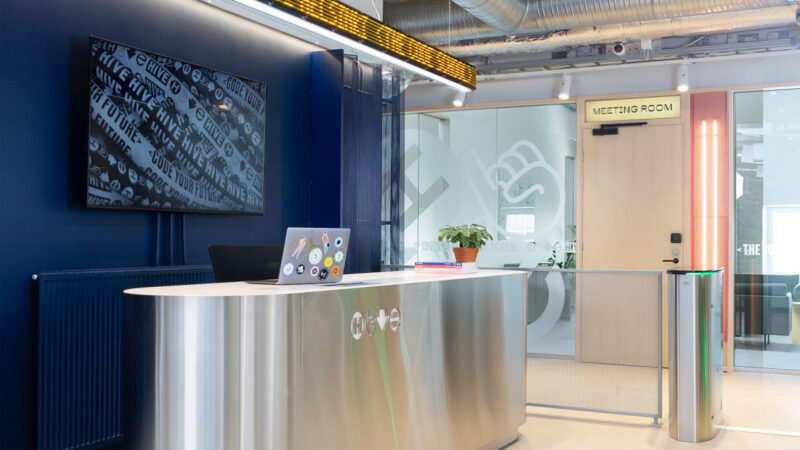 The modern reception area of Hive Helsinki, featuring a sleek, metallic desk with a laptop and a potted plant. On the left, a large screen displays "HIVE CODE YOUR FUTURE" in repeated text. To the right, a wooden door leads to a "MEETING ROOM," and a turnstile entrance is visible, depicting the welcoming and functional entrance to the coding school in Helsinki, Finland.
