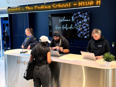 The reception desk at Hive Helsinki, featuring staff members assisting visitors or students. A large screen behind the desk displays "Welcome to Hive Helsinki," with a digital ticker board above it. This vibrant space at the Helsinki campus welcomes individuals into our unique coding education environment.