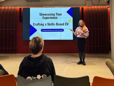 A woman named Lumi Tomperi is giving a presentation in a modern event space at Hive Helsinki. She stands beside a large screen that displays the title "Showcasing Your Experience: Crafting a Skills-Based CV" and "A Practical Workshop for Career Changers." An audience member, seen from behind, is seated in the foreground.