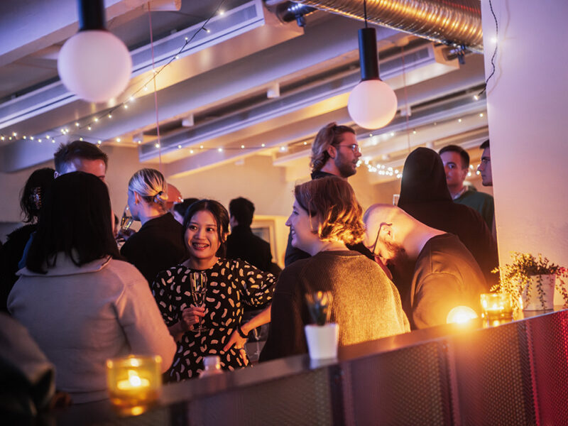 A lively social gathering at Hive Helsinki, with groups of people mingling and chatting under soft, festive lighting. A woman with dark hair in a patterned dress is smiling in the center, holding a drink. The warm ambiance suggests a relaxed and welcoming community environment in Helsinki.