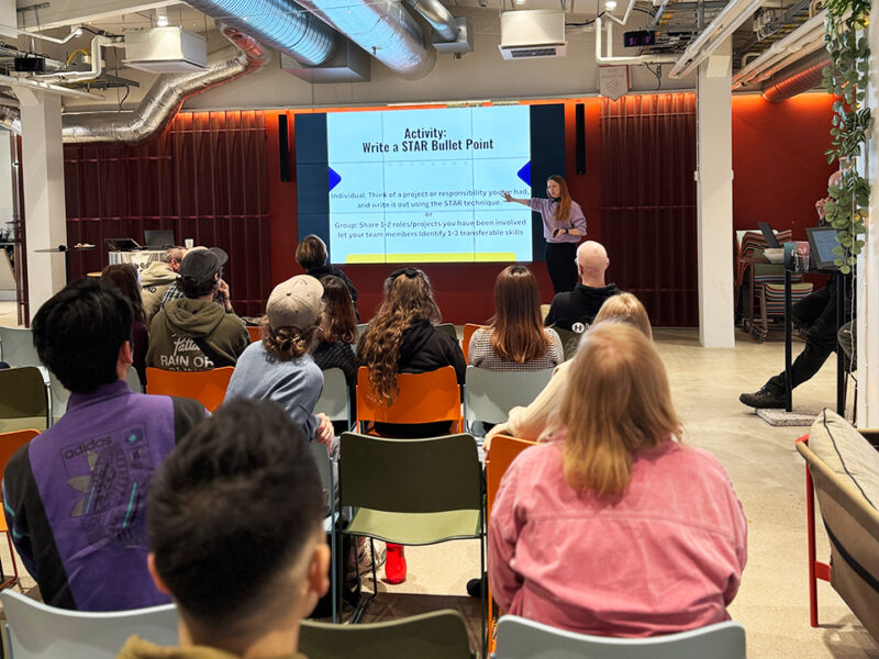 A presenter at Hive Helsinki conducting a workshop on "Showcasing Your Experience: Crafting a Skills-Based CV," specifically on the "Activity: Write a STAR Bullet Point." An audience is seated, facing the screen, actively engaged in the session. The room features an industrial design with exposed ductwork and red textured walls.