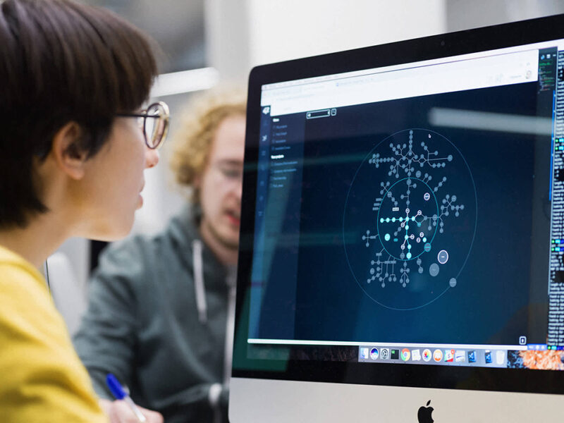 A Hive Helsinki student, wearing glasses and a yellow shirt, intently viewing a complex data visualization or network graph on an Apple iMac screen. Another student is blurred in the background, highlighting the focused and in-depth learning experiences in coding and tech at the Helsinki campus.
