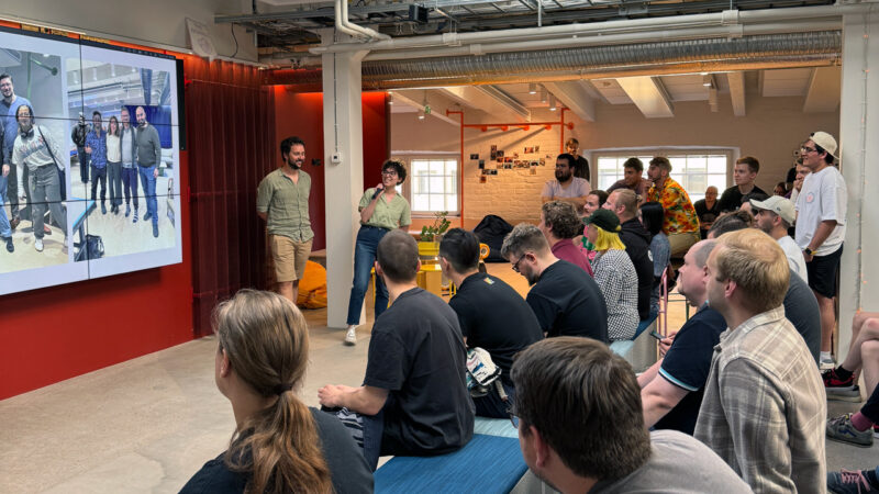 Two Hive Helsinki students presenting to a large, engaged audience in a modern event space. A large screen behind them displays images, while the audience listens intently. This scene highlights the practical presentation skills and collaborative learning environment at the coding school in Helsinki, Finland.