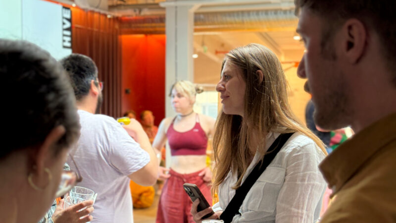 A Hive Helsinki student or community member, smiling and engaging in conversation at a social event. She is holding a smartphone, with other people and elements of the modern Hive Helsinki campus visible in the background, showcasing the vibrant networking and community life in Helsinki, Finland.