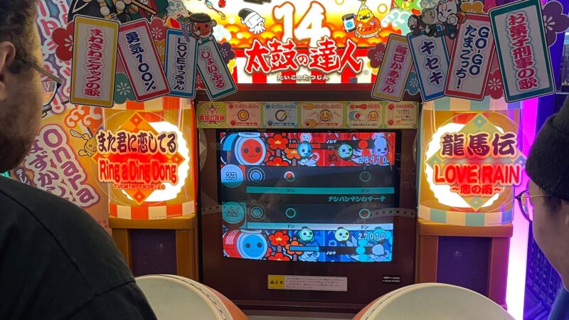 Two individuals focused on playing a Japanese drum arcade game at the Assembly event in Helsinki. The brightly lit screen displays game elements and scores, while various colorful signs are visible around the machine. This captures the fun and interactive side of gaming culture that appeals to Hive Helsinki students.