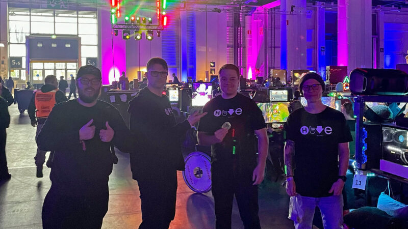 Four smiling individuals, wearing Hive Helsinki branded t-shirts, pose together at the Assembly event in Helsinki. They give thumbs-up and appear friendly, representing Hive Helsinki's presence and engagement within Finland's tech and gaming communities. The background shows many gaming setups under colorful lights.